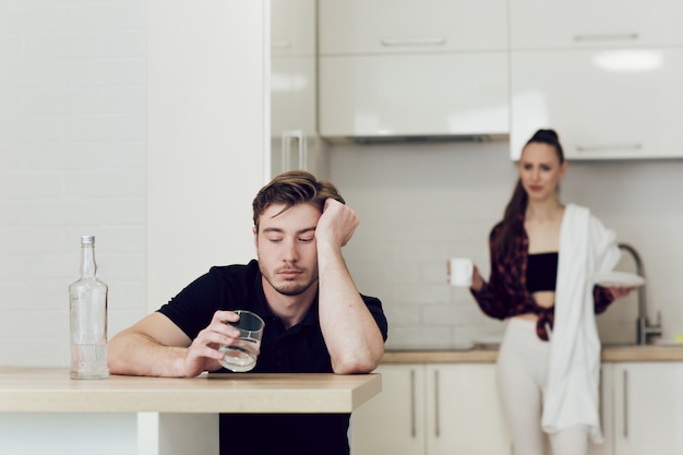 Ein Mann trinkt, während er an einem Tisch in der Küche sitzt, eine Frau hinter ihm streitet und schreit.