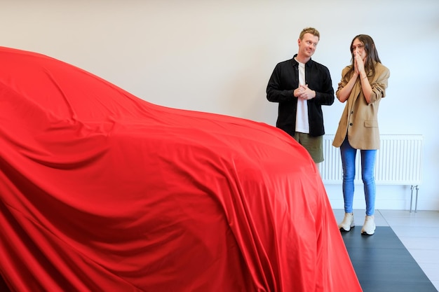 Ein Mann überrascht seine geliebte Freundin, indem er ihr ein neues Auto kauft Ein modernes Auto steht unter einem roten Tuch in einem Autohaus