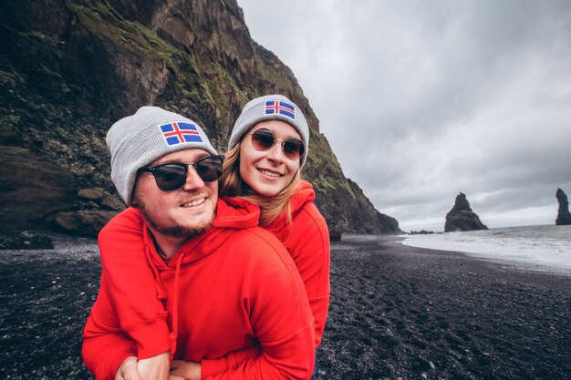 Ein Mann und ein Mädchen in roten Pullovern und grauen Hüten umarmen sich an einem schwarzen Strand neben Vic