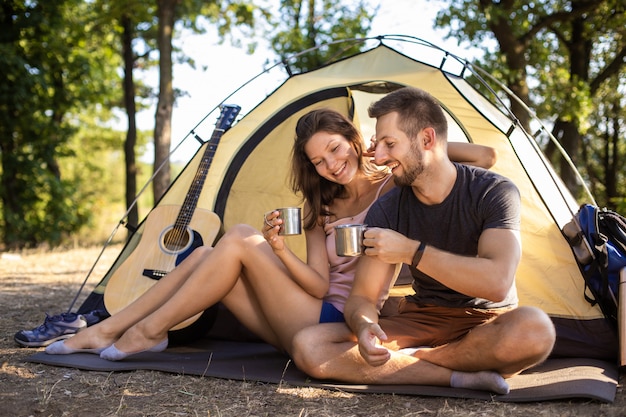 Ein Mann und eine Frau auf einem Campingausflug trinken Tee, während sie in der Nähe eines Zeltes sitzen.