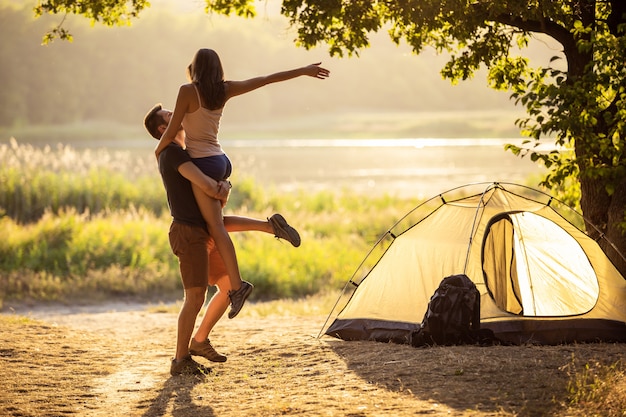 Ein Mann und eine Frau bei einer Wanderung mit Rucksäcken in der Nähe eines Zeltes bei Sonnenuntergang umarmen sich.