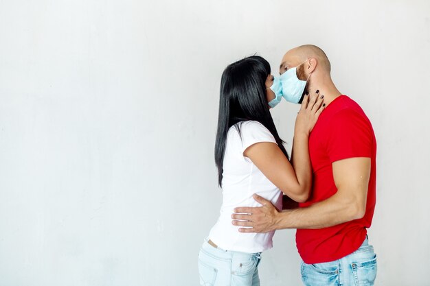 Ein Mann und eine Frau in medizinischen Masken küssen sich gegen eine weiße Wand und zeigen Einschränkungen des Coronavirus