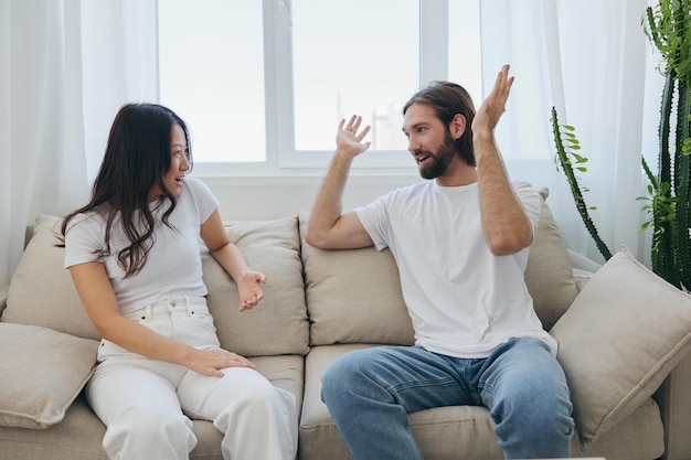 Ein Mann und eine Frau sitzen zu Hause auf der Couch in weißen, stylischen T-Shirts und unterhalten sich fröhlich lächelnd und lachend zu Hause Freundschaft zwischen Männern und Frauen