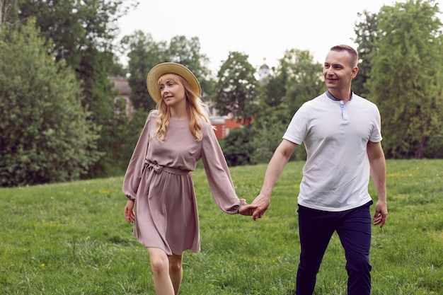 Ein Mann und eine verliebte Frau in Kleid und Hut gehen im Sommer auf einer grünen Wiese unter einem Baum spazieren