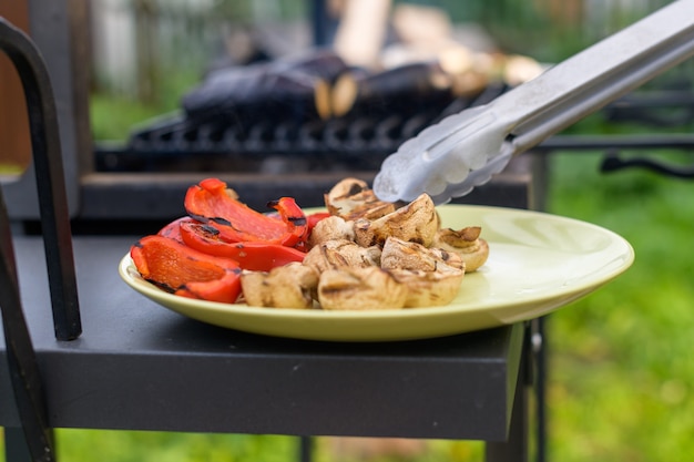 Ein Mann verteilt gegrillte Paprika und Pilze auf einer runden Keramikplatte mit einer Küchenzange