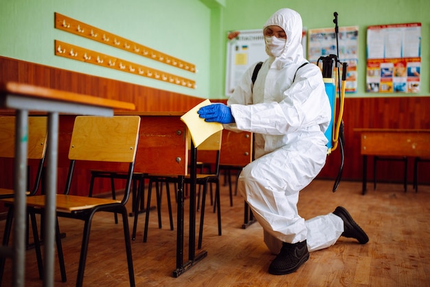 Ein Mann von der Desinfektionsgruppe räumt mit einem gelben Lappen den Schreibtisch in der Schule auf. Professioneller Arbeiter sterilisiert das Klassenzimmer, um die Ausbreitung von Covid-19 zu verhindern. Gesundheitswesen von Schülern und Studenten Konzept.