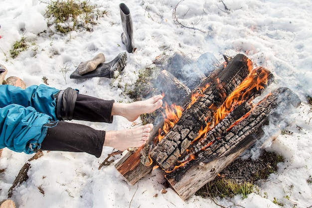 Ein Mann wärmt seine barfüßigen Füße in der Nähe des Feuers im Kiefernwald, der im Winter mit Schnee bedeckt ist