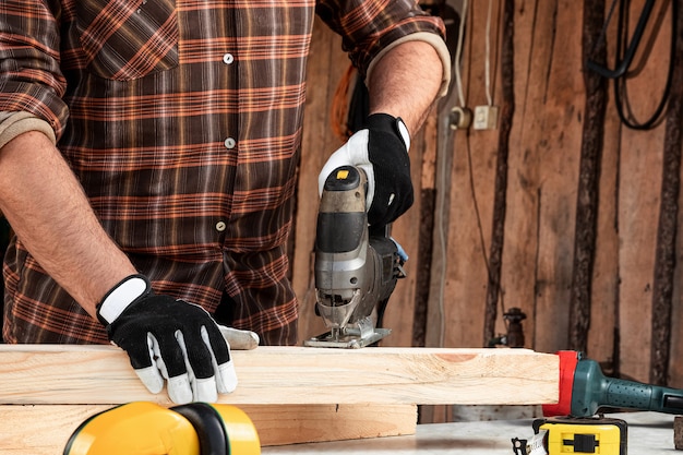 Ein Manntischler schneidet einen Holzbalken unter Verwendung einer elektrischen Laubsäge, männliche Hände mit einer elektrischen Laubsägennahaufnahme.