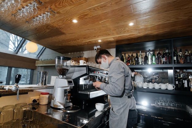 Ein maskierter Barista bereitet an der Bar in einem Café einen exquisiten, köstlichen Kaffee zu Die Arbeit von Restaurants und Cafés während der Pandemie