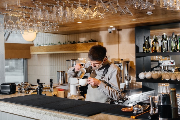 Ein maskierter barista bereitet an der bar in einem café köstlichen kaffee zu. die arbeit von restaurants und cafés während der pandemie.