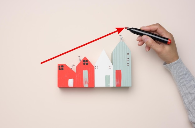 Ein Miniatur-Holzhaus und die Hand einer Frau zeichnen ein Diagramm mit wachsenden Indikatoren