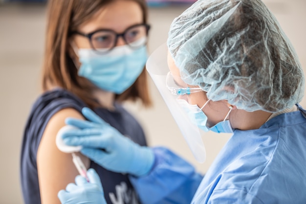 Ein Mitarbeiter von Helathcare, der Handschuhe, Schutzschild und eine Kappe trägt, trägt den Impfstoff aus einer Spritze in den Arm eines jungen Patienten auf.