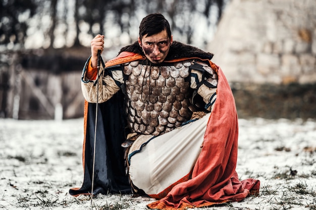 Ein mittelalterlicher Krieger in Kettenrüstung und Mantel kniet nieder und hält nach dem Kampf seine Hand am Schwert.