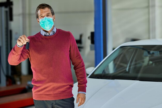 Ein mittlerer erwachsener Mann, der eine schützende Gesichtsmaske trägt, während er die Schlüssel seines reparierten Autos in einer Werkstatt hält