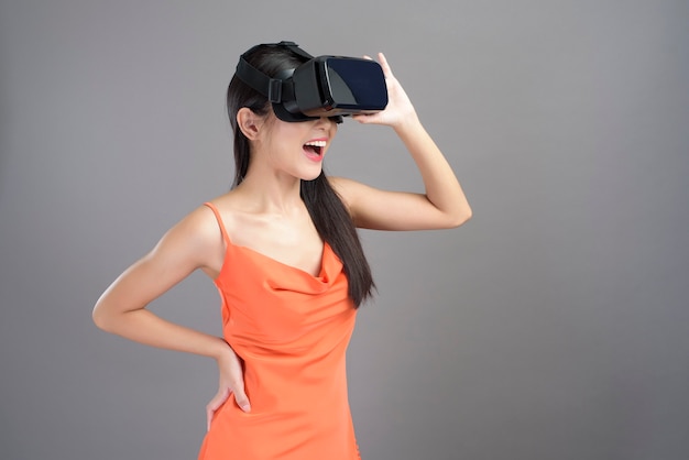 Ein Modeporträt der schönen Frau im orangefarbenen Kleid trägt eine auf Grau isolierte Virtual-Reality-Box.