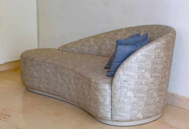 Foto ein modernes sofa mit einer einzigartigen gekrümmten form liegt an einer weißen wand und der boden ist gepflastert. es ist in einem hellfarbenen stoff mit einer ausgeprägten textur gepolstert. ein blaues kissen ruht auf dem sofa.