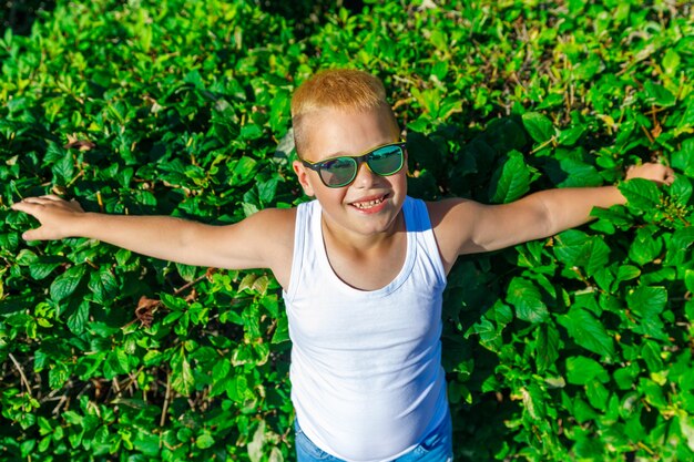 Ein modischer Junge in weißem T-Shirt und schwarzer Brille steht vor einem Hintergrund grüner Büsche