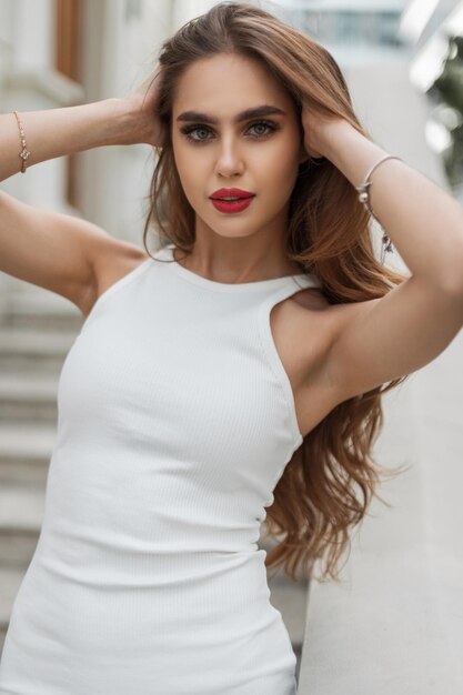 Ein modisches junges Schönheitsmodell mit roten Lippen in einem eleganten weißen Kleid steht im Freien und repariert ihre Haare