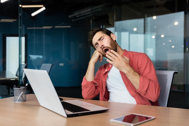 Ein müder junger Mann in einem roten Hemd sitzt am Schreibtisch im Büro und gähnt und bedeckt seinen Mund mit