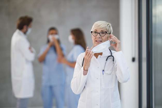 Ein müder leitender Arzt, der während der Covid-19-Pandemie in einem Krankenhausflur eine kurze Pause einlegt und eine Schutzmaske hält. Blick in die Kamera.