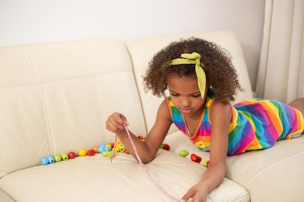 Foto ein mulatt-kind auf der couch konzentriert sich auf die perlen einer halskette gelbes kopfband gestreiftes regenbogenkleid ein moment der kreativität und des lernens zu hause bildung und therapie mit spielzeug