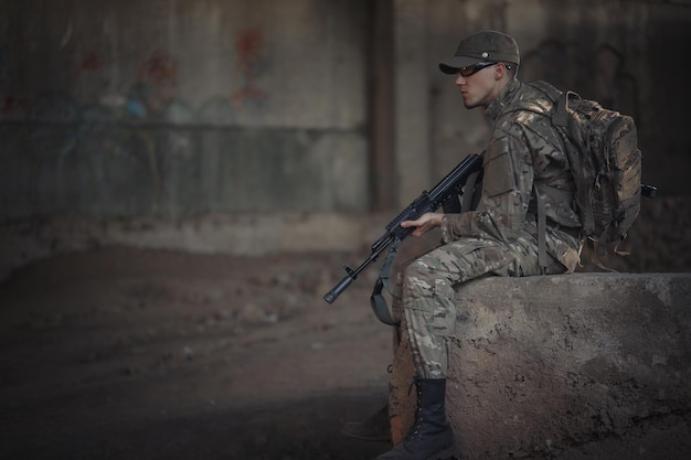 Ein mutiger ukrainischer Soldat mit Rucksack auf dem Rücken in grauer Tarnuniform mit taktischer Brille und Mütze mit Maschinengewehr in der Hand sitzt in einem zerstörten Gebäude