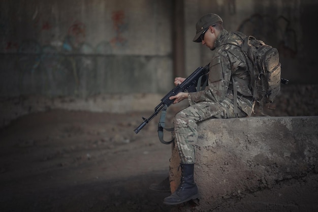 Ein mutiger ukrainischer Soldat mit Rucksack auf dem Rücken in grauer Tarnuniform mit taktischer Brille und Mütze mit Maschinengewehr in der Hand sitzt in einem zerstörten Gebäude