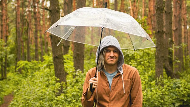 Ein nachdenklicher Mann mit Regenschirm in der Hand im Wald Ein Hipster mit Dreadlocks in Freizeitkleidung versteckt sich vor dem Regen unter einem Regenschirm im Wald