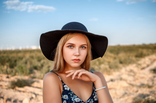 Ein Nahaufnahmeporträt blauäugigen Blondine in einem schwarzen Hut und in einem Blumendruckkleid Sie wirft in einem Weinberg während des Sonnenuntergangs auf