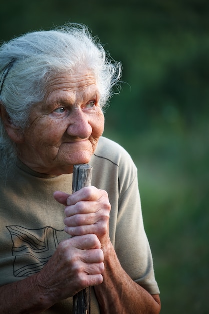 Ein Nahaufnahmeporträt einer alten Frau mit dem grauen Haar oben lächelnd und schauend, ihr Kinn auf einem Stock stillstehend, als ob gehend mit einem Stock, Gesicht in den tiefen Falten