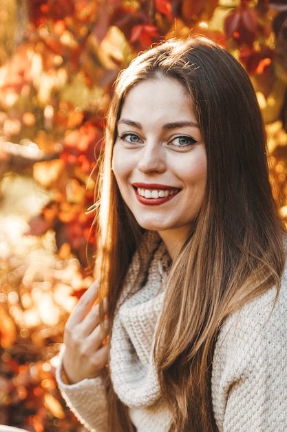 Ein Nahaufnahmeporträt einer Frau auf dem Hintergrund der roten und gelben Blätter im Park. Sie trägt einen beigen Pullover und einen roten Lippenstift. Sie lächelt und ihr Haar ist lang.