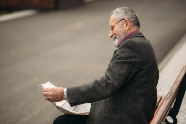 Ein netter Großvater mit einem schönen Bart in einer grauen Jacke sitzt auf einer Bank im Park und liest eine Zeitung