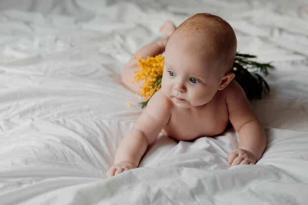 Ein neugeborenes Baby, das mit einem Mimosenzweig bedeckt ist, liegt auf seinem Bauch auf einem weißen Bett