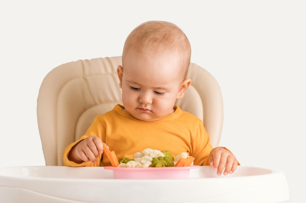 Ein Neugeborenes im Alter von einem Jahr probiert zum ersten Mal gekochtes Gemüse. Ein Baby nimmt vorsichtig eine gekochte Karotte, während es auf einem Fütterungsstuhl auf weißem Hintergrund sitzt