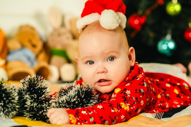 Ein neugeborenes Junge mit einem roten Weihnachtsmannshut liegt auf dem Bett in der Nähe des Weihnachtenbaums