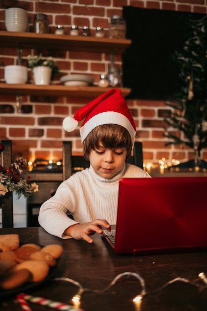 Ein Neujahrskind in einem weißen Pullover und einer roten Neujahrsmütze sitzt an einem Tisch und tippt einen Brief in einen roten Laptop