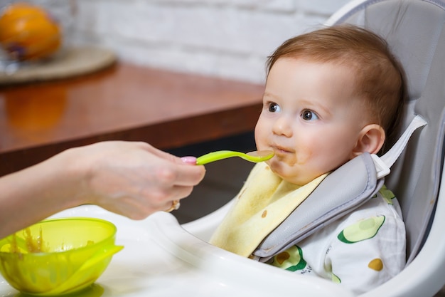 Ein neun Monate altes lächelndes Baby sitzt an einem weißen Tisch in einem Hochstuhl und isst mit einem Löffel aus einer Schüssel. Mama füttert das Baby mit einem Löffel. Unscharfer Hintergrund. Gesundes Essen für Kinder. Kinder essen.