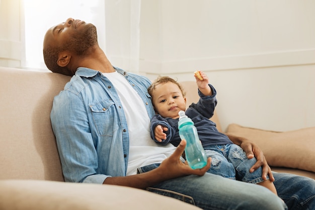 Ein Nickerchen machen. Dunkelhaariger müder afroamerikanischer Mann, der schläft und seinen kleinen Sohn und seine Flasche hält und auf der Couch sitzt