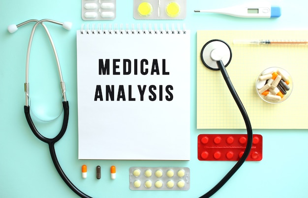 Ein Notizbuch mit dem Text MEDIZINISCHE ANALYSE liegt ordentlich gefaltet zwischen den Pillen, einem Stethoskop und einem gelben Notizbuch. Medizinisches Konzept