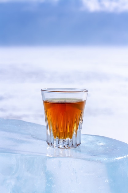 Ein orangefarbenes alkoholisches Getränk in einem Glas steht auf einem Eisstück. Eis ist in der Sonne geschmolzen. Heller unscharfer Hintergrund. Platz kopieren. Seitenansicht. Vertikal.