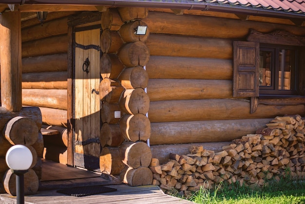 Ein ordentlicher großer Holzstapel vor dem Hintergrund eines Holzhauses aus Baumstämmen mit einem kleinen Fenster. Sonniger Frühlings- oder Sommertag