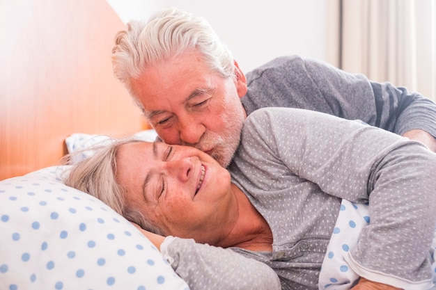 Ein paar ältere Männer und Frauen, die aufwachen und mit einer Umarmung lächeln, während sie zu Hause im Bett sind. Er küsst sie mit Liebe für ein gemeinsames Leben