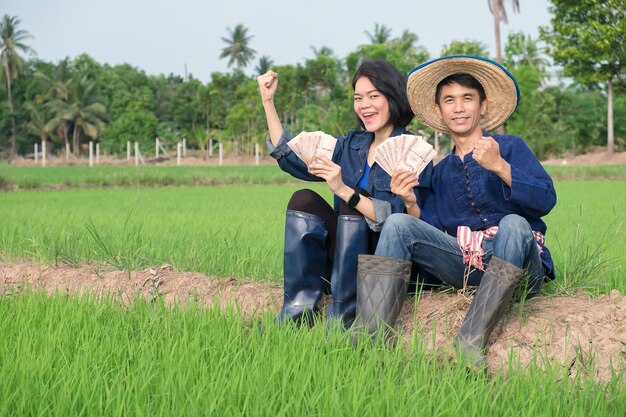 Ein Paar asiatischer Bauern und Bäuerinnen in traditionellen Kostümen halten glücklich Banknoten und heben ihre Hände auf einem Feld.