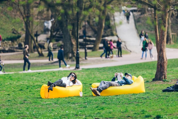 Ein Paar, das auf einer gelben aufblasbaren Matratze im Stadtpark liegt, liest ein Buch und surft am Telefon