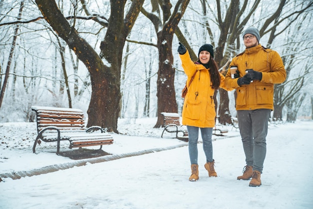 Ein Paar, das durch den verschneiten Stadtpark geht und Kaffee trinkt, um etwas geradeaus zu zeigen