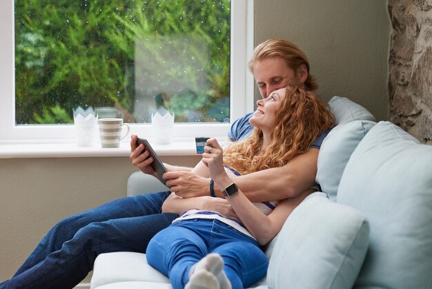 Ein Paar entspannt sich zusammen auf der Couch und verwendet eine Kreditkarte, um Online-Einkäufe zu tätigen Ein Paar entspannt sich zusammen auf der Couch und verwendet eine Kreditkarte, um Online-Einkäufe zu tätigen