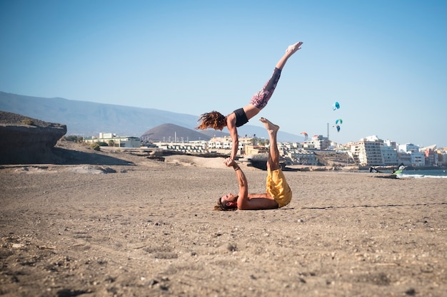 Ein paar Erwachsene, die schweigend Yoga oder Acroyoga am Strand machen - Frau und Mann konzentrieren sich auf die Zusammenarbeit - Stadt im Hintergrund