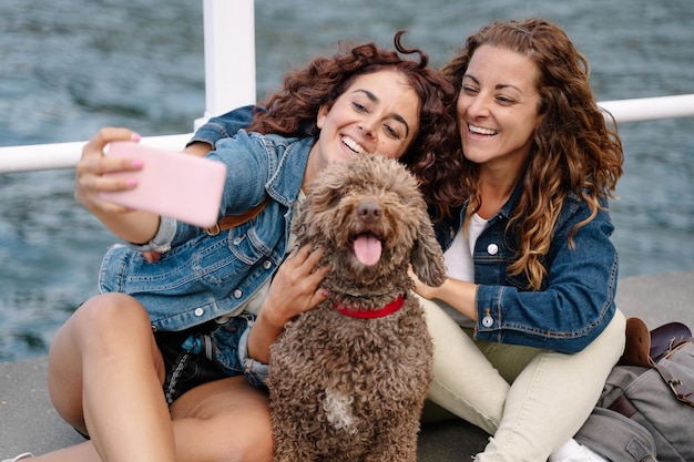 Ein paar Freunde, die ein Foto mit braunem Wasserhund machen. Horizontale Ansicht von Frauen, die mit Haustier reisen. Lifestyle und Technologie mit Tieren im Freien.