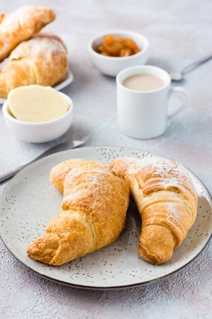 Ein paar frische knusprige Croissants auf einem Teller, eine Tasse Kaffee und Schüsseln mit Marmelade und Butter auf einem hell strukturierten Hintergrund. Kaffeepause oder Frühstück. Vertikale Ansicht