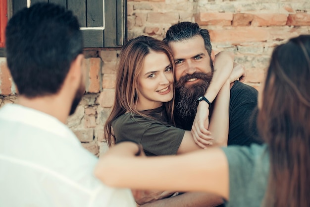 Ein Paar hübscher bärtiger Mann mit langem Bart und stylischem Haar und eine blonde, hübsche, sexy Frau oder ein Mädchen, die ihre Freunde am sonnigen Sommertag auf steinernem Wandhintergrund umarmen und anlächeln